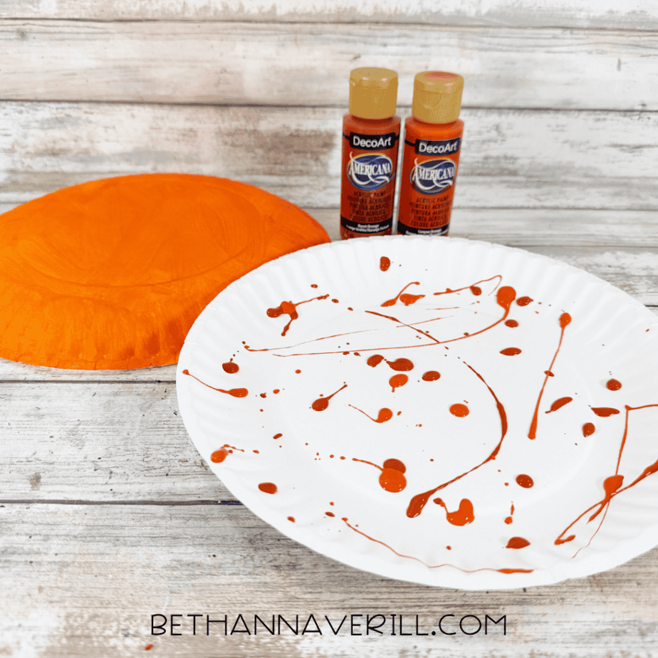 White paper plate covered with scattered orange and brown acrylic paint droplets for a fall leaf painting activity.