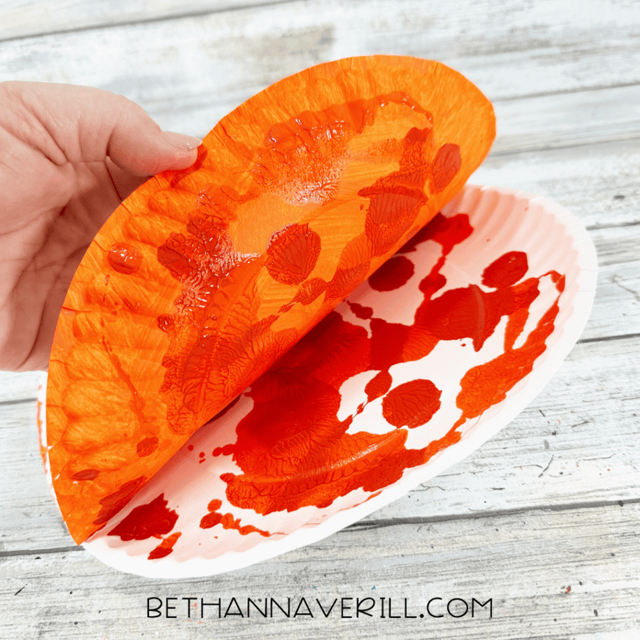 Hand lifting an orange painted paper plate to reveal textured paint patterns created by pressing two plates together.