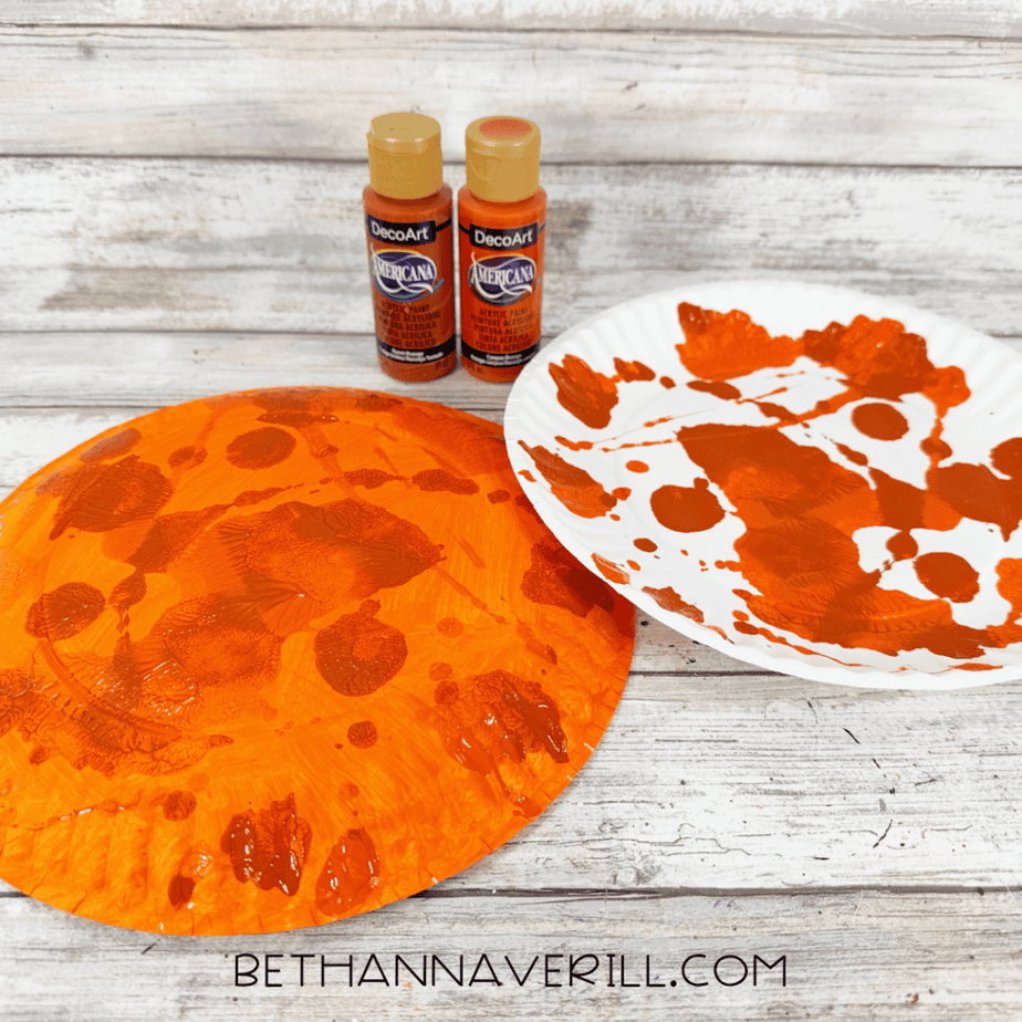 Orange painted paper plate with darker orange and brown textured paint patterns beside a white plate covered in paint.
