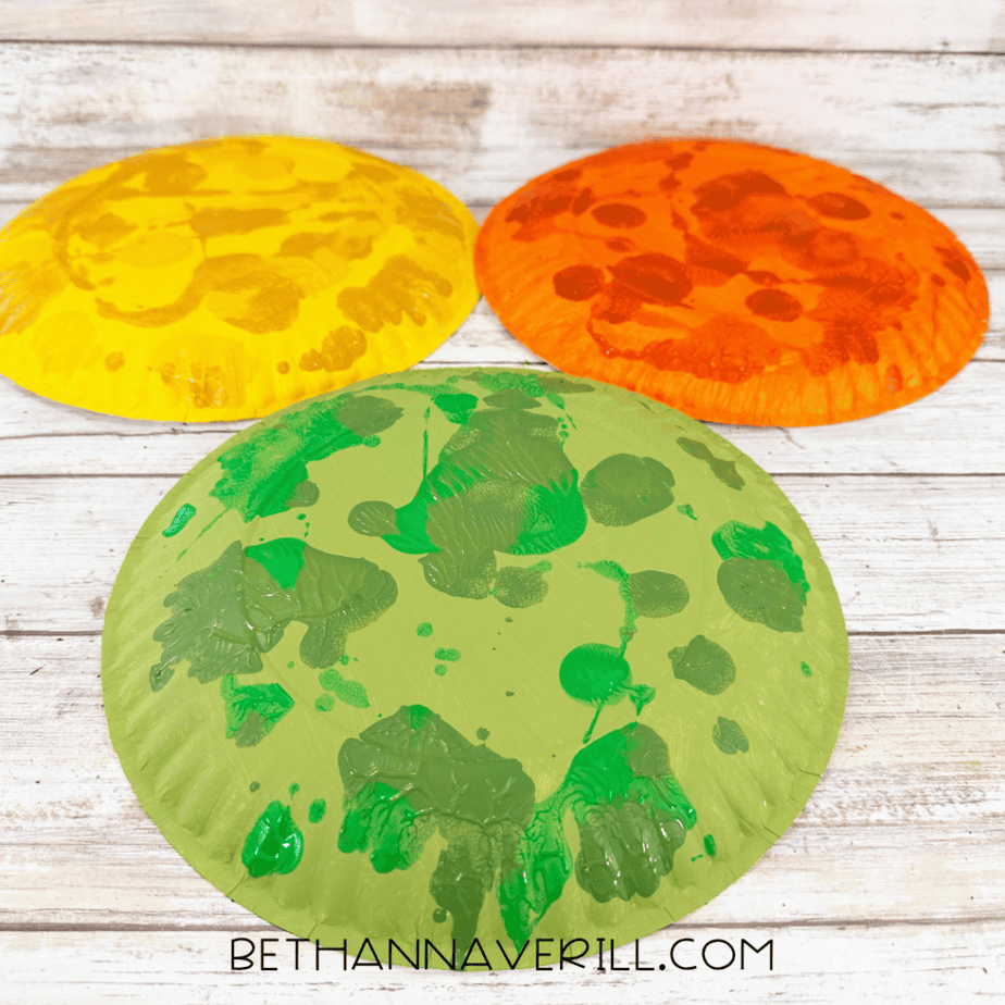 Three painted paper plates in yellow, orange, and green decorated with contrasting paint patterns for autumn leaf crafts.