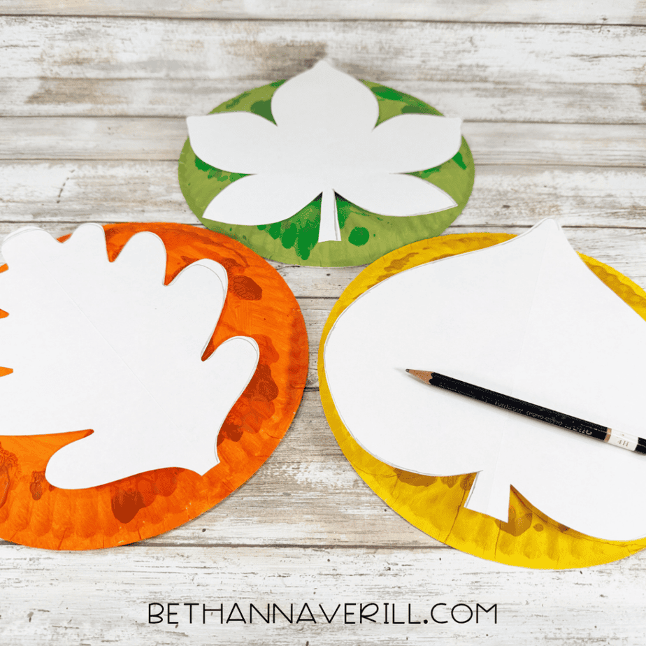 White leaf-shaped paper templates placed over green, orange, and yellow painted paper plates with a pencil for tracing.