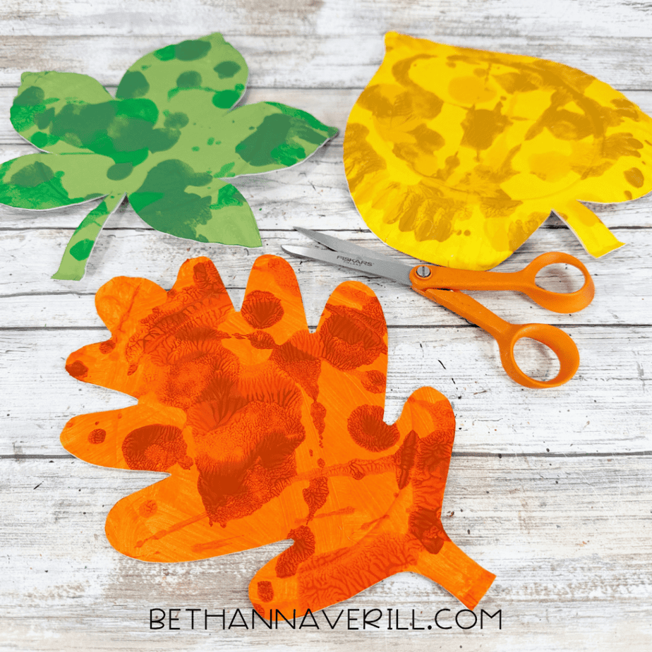 Three autumn leaf shapes cut from painted paper plates, including an orange oak leaf, yellow leaf, and green maple-shaped leaf.