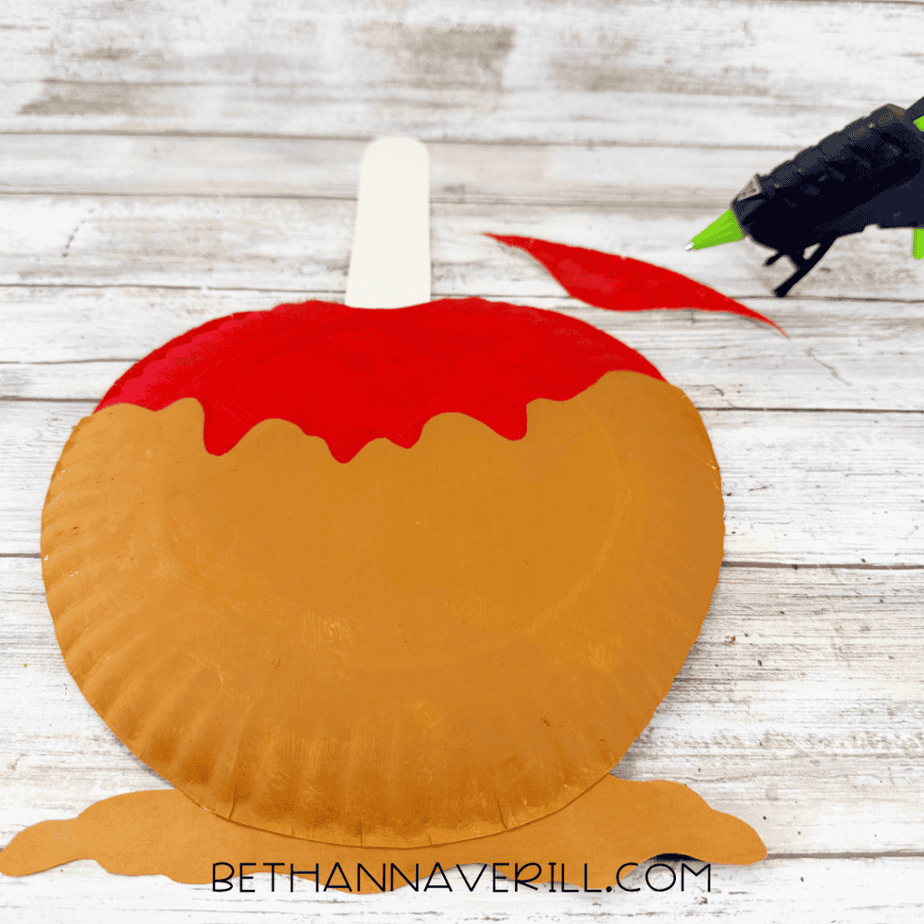 Caramel apple paper plate craft with a white cardstock stick attached at the top and a brown cardstock piece beneath the apple.