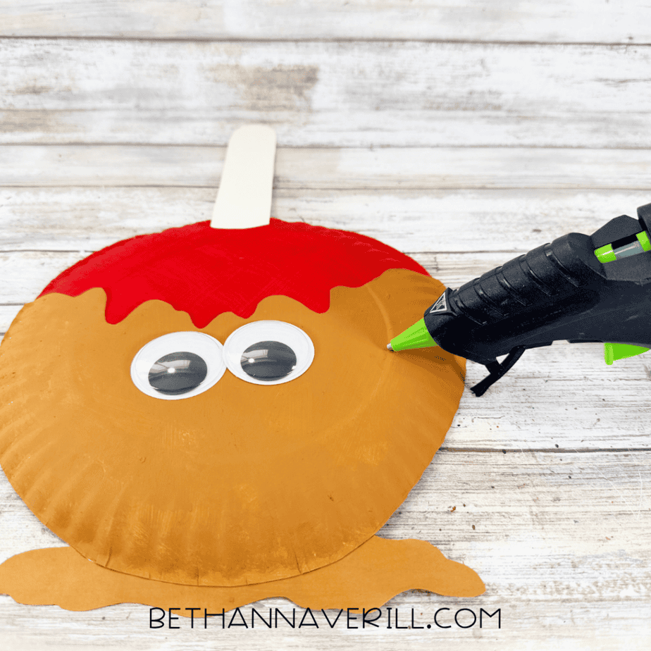 Two large googly eyes being attached to a caramel apple paper plate craft with a hot glue gun nearby.