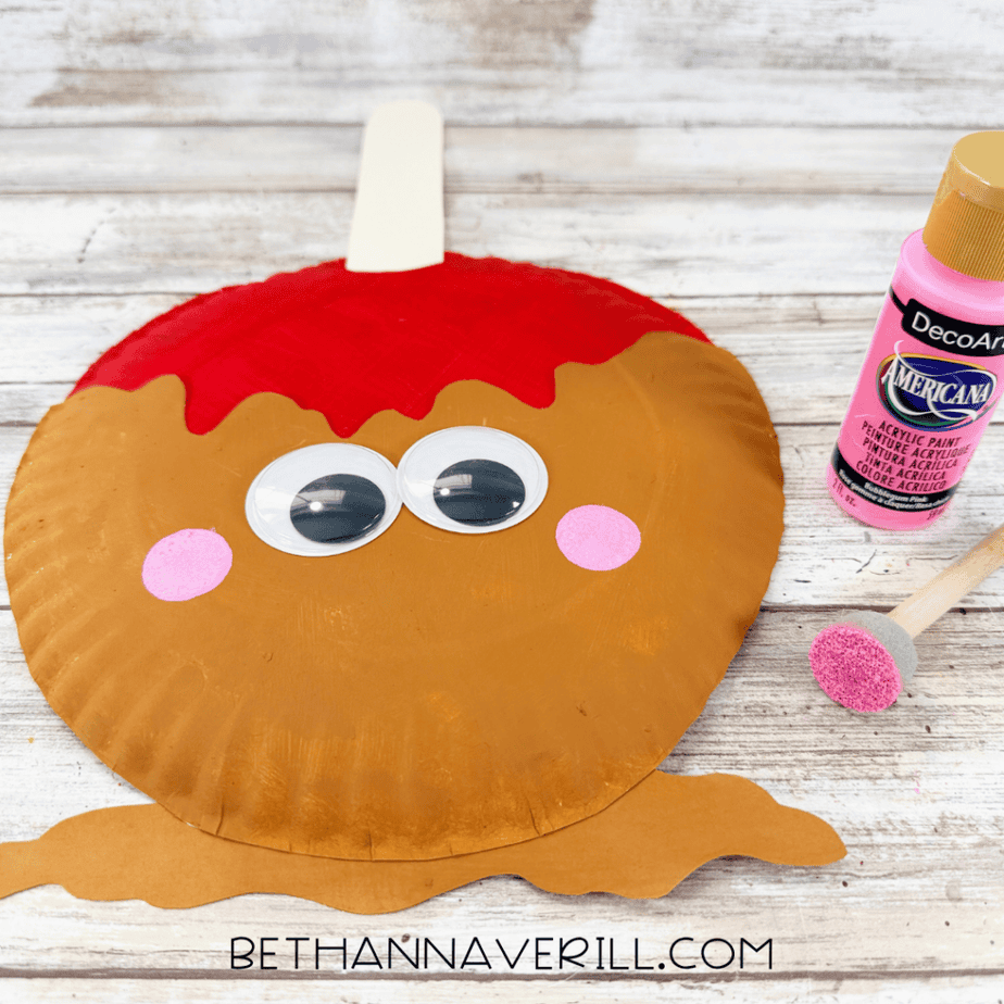 Caramel apple paper plate craft with googly eyes and pink painted cheeks beside a pink acrylic paint bottle and sponge brush.
