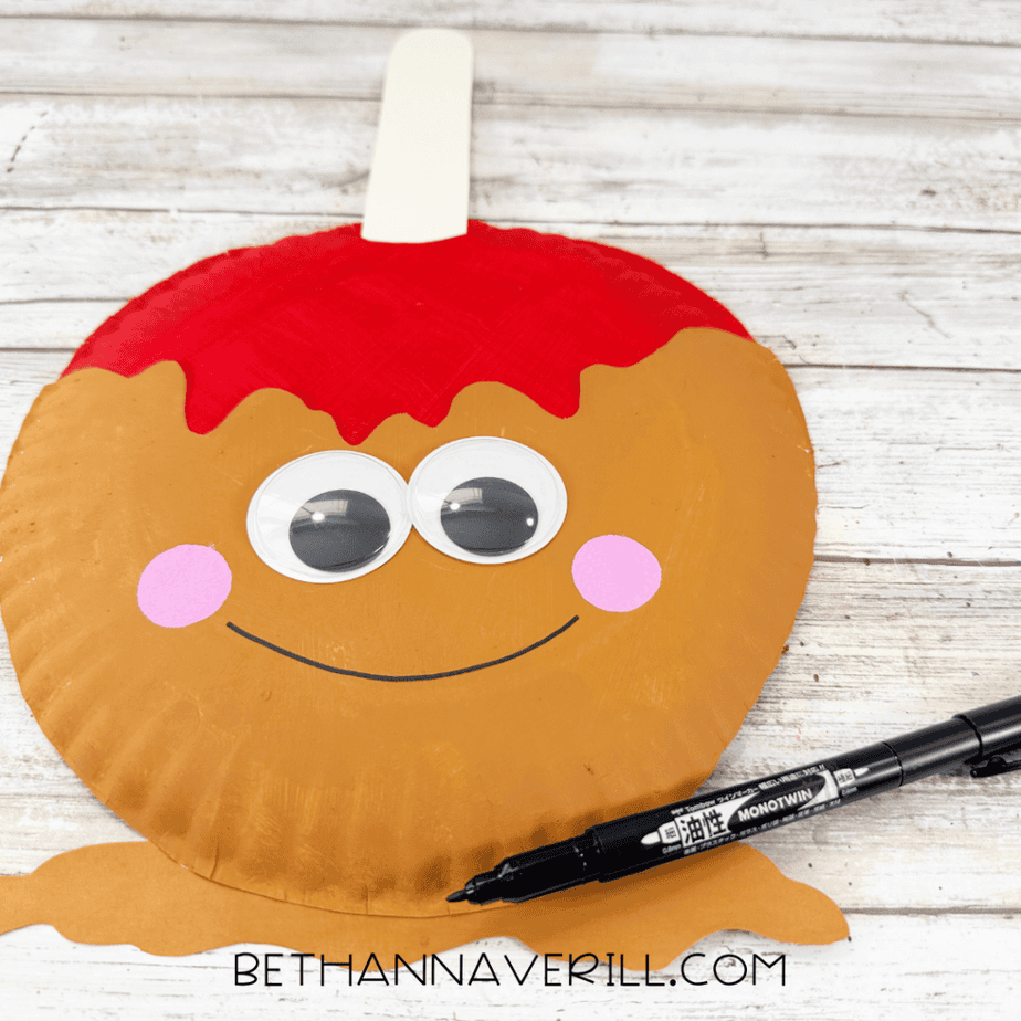 Smiling caramel apple paper plate craft with large googly eyes, pink cheeks, and a black marker beside it.