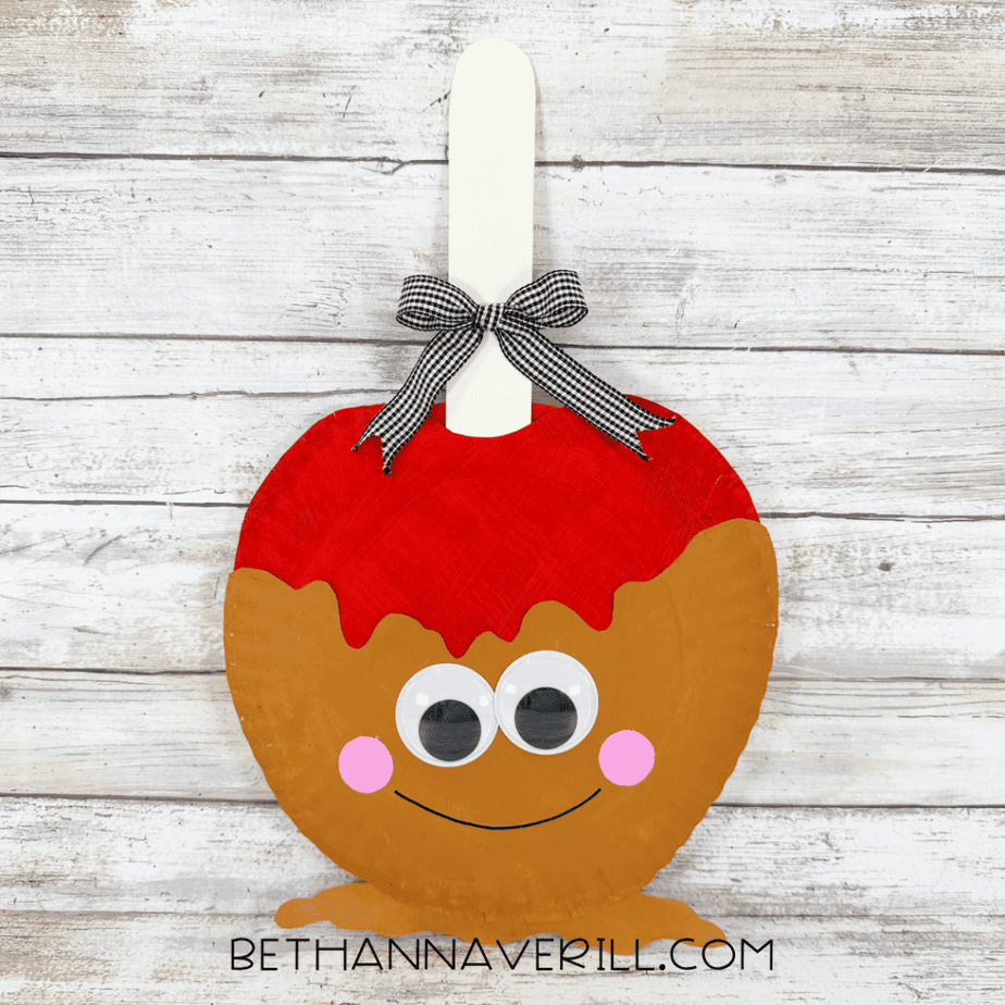 Finished caramel apple paper plate craft with red topping, caramel brown body, googly eyes, pink cheeks, a smiling face, and a gingham ribbon bow.