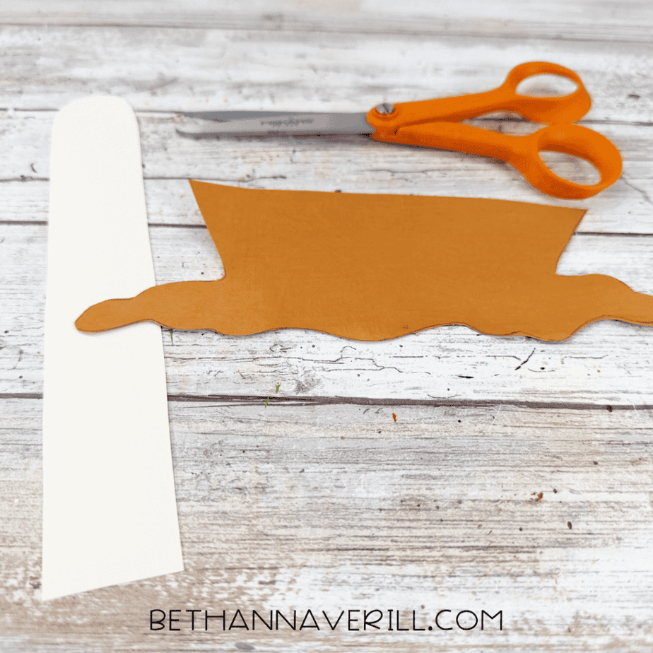 Brown cardstock caramel drip cutout and a long white cardstock strip beside orange scissors.