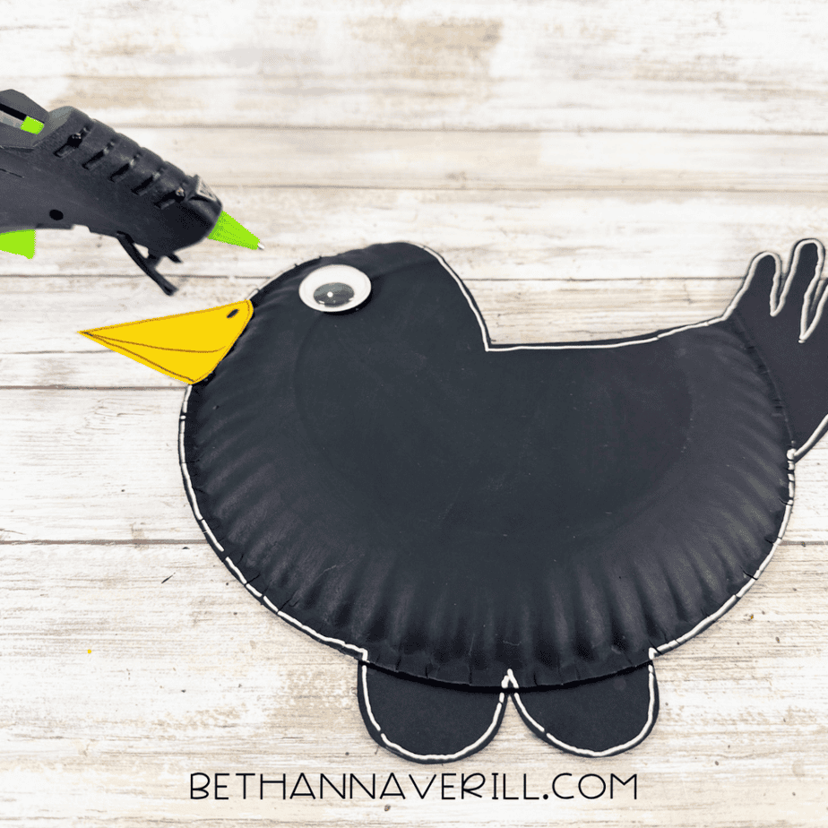 Black paper plate crow with a yellow beak, googly eye, white outline, tail, and black feet being assembled with a hot glue gun.
