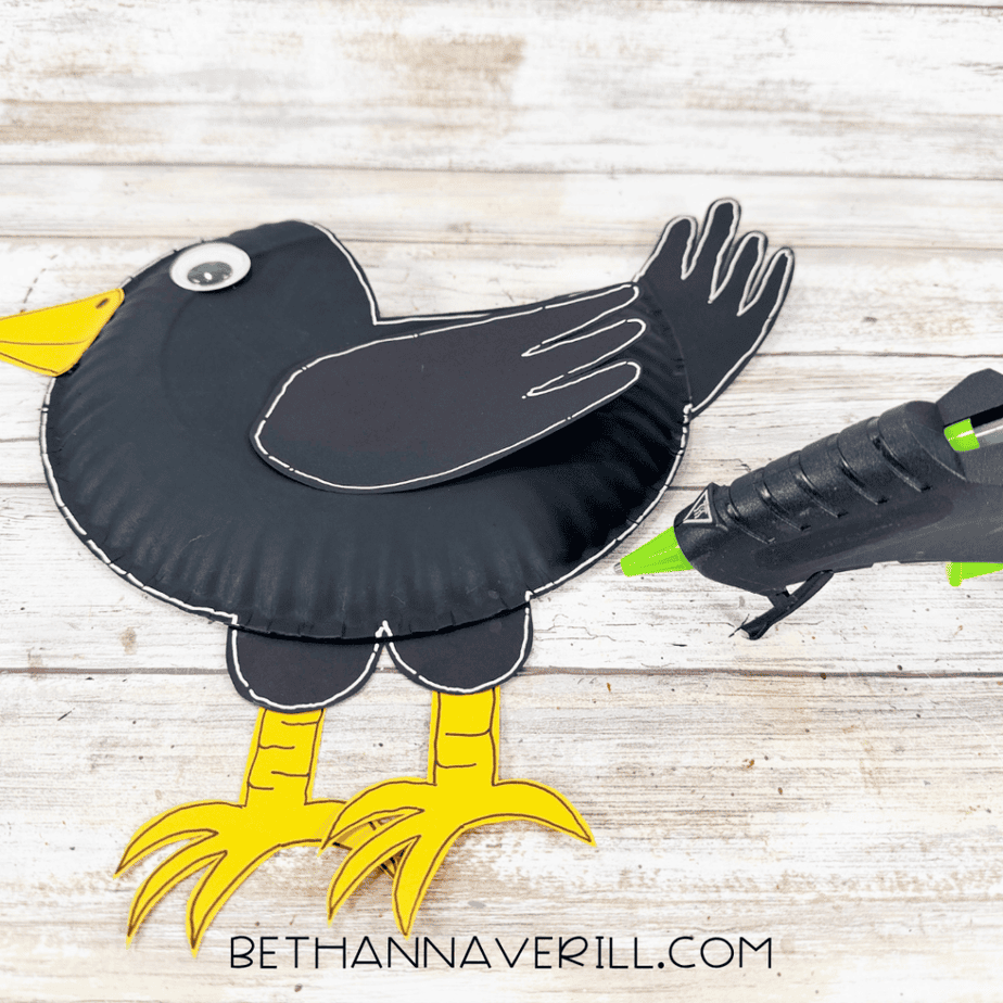 Black paper plate crow with black wing, tail, yellow beak, yellow legs and feet, googly eye, and white outline details.