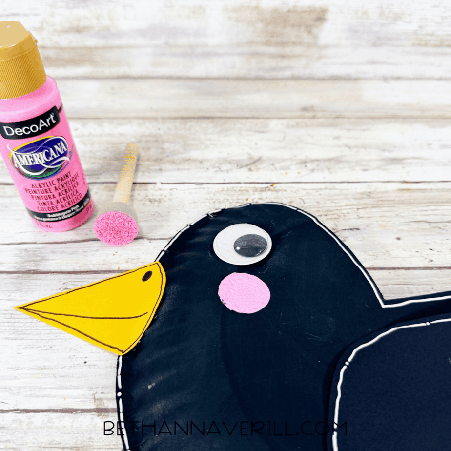 Close-up of the crow craft with a pink painted cheek beside pink acrylic paint and a sponge dauber.