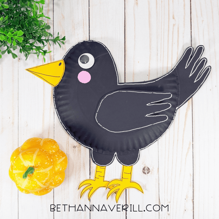 Finished black paper plate crow with yellow beak and feet, pink cheek, googly eye, black wing, and white outline displayed beside a small yellow pumpkin.