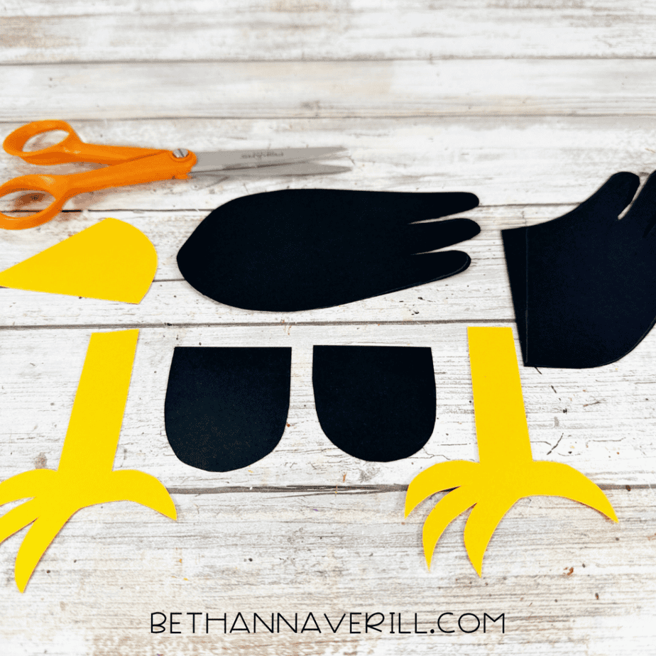 Cut black paper wings and body pieces with yellow paper legs, feet, and beak arranged on a white wood surface.