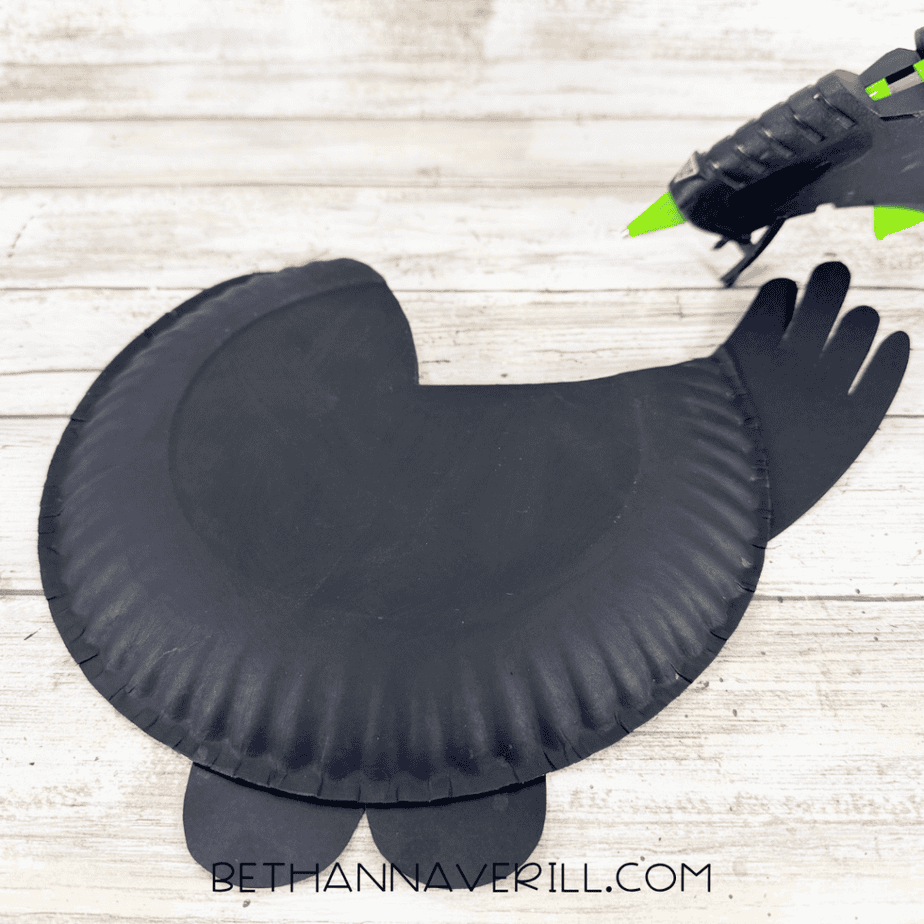 Black paper plate bird body with black tail and feet pieces attached while a hot glue gun rests nearby.