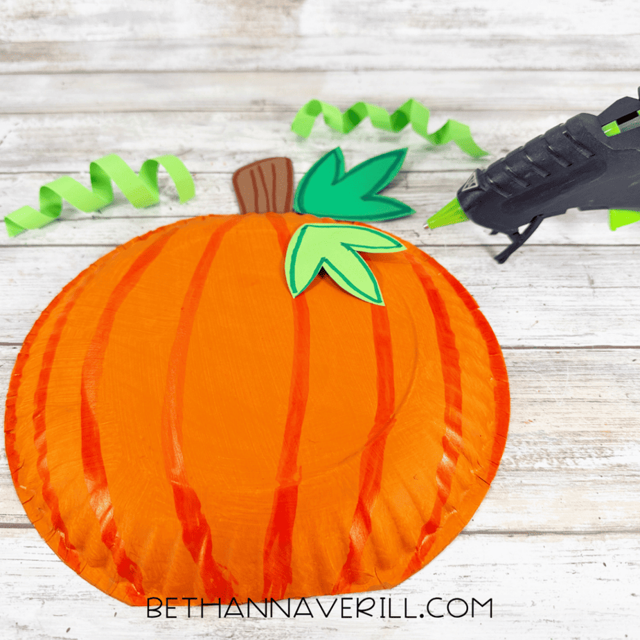 Orange paper plate pumpkin with a brown stem, green leaves, curled green paper tendrils, and a hot glue gun nearby.