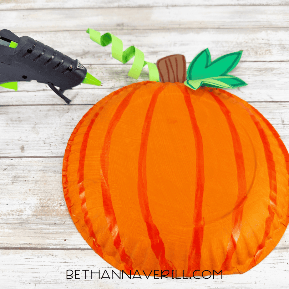 Orange paper plate pumpkin with a brown stem, green leaves, curled green paper tendrils, and a hot glue gun nearby.