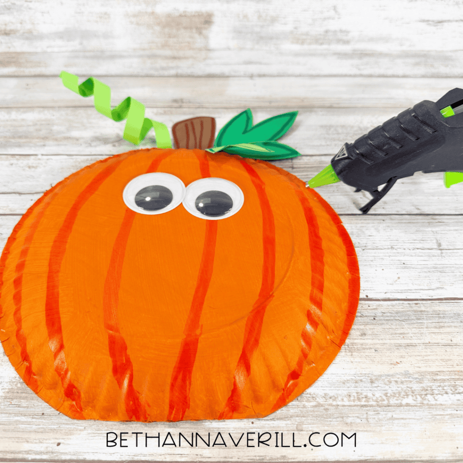 Orange paper plate pumpkin with stem, leaves, and curled paper vine, shown while large googly eyes are being added.