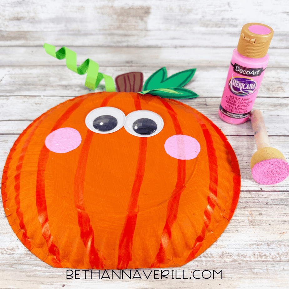 Orange paper plate pumpkin with large googly eyes and two round pink painted cheeks, with pink paint and a sponge applicator beside it.