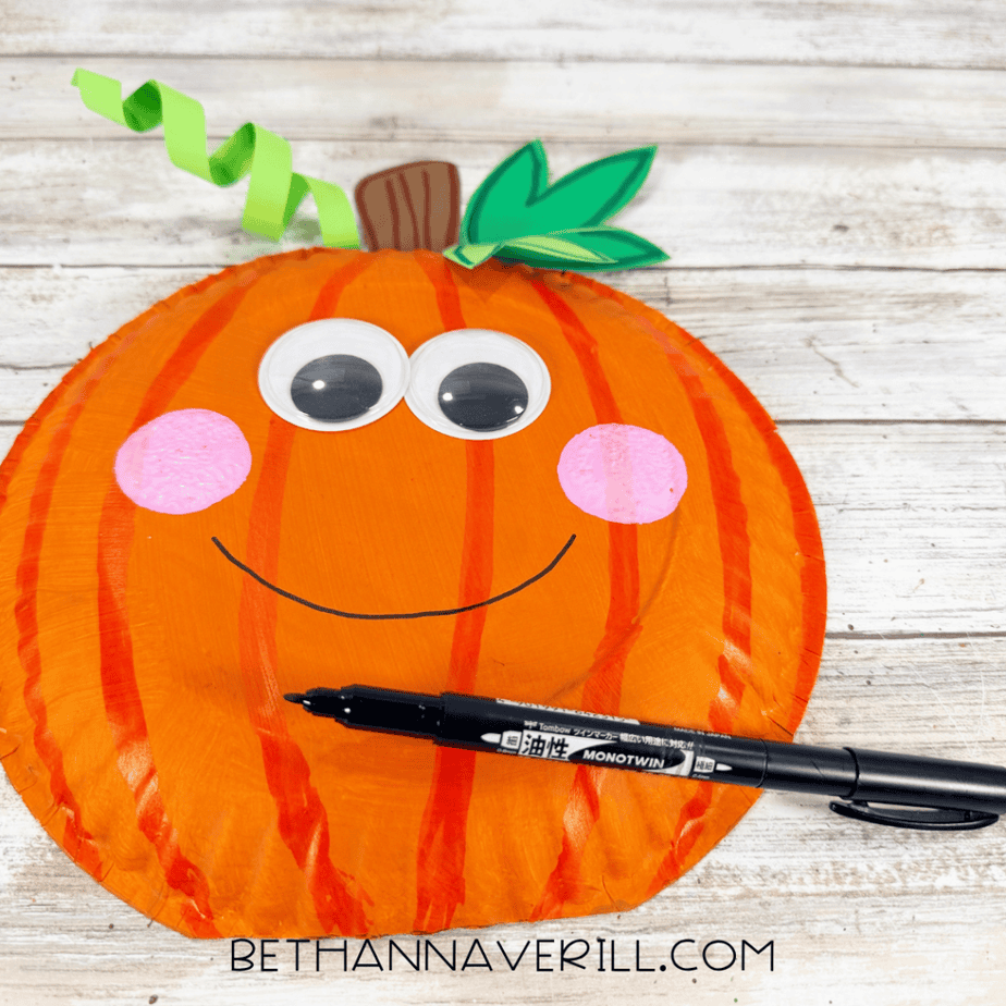 Paper plate pumpkin with googly eyes, pink cheeks, green leaves, curled vine, and a curved smile being drawn with a black marker.