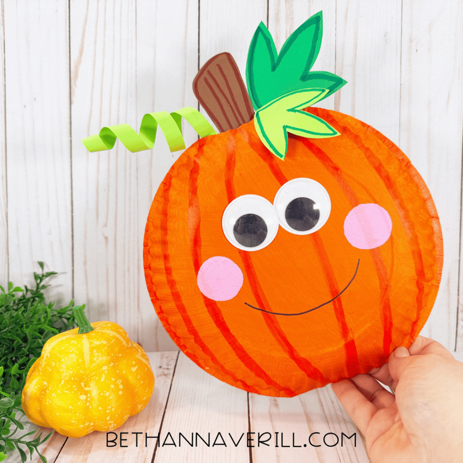 Finished smiling orange paper plate pumpkin craft held upright, with large googly eyes, pink cheeks, green leaves, a brown stem, and curled green vine.