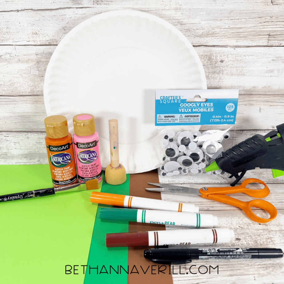 Paper plate craft supplies arranged on a white wood surface, including paper plates, orange and pink paint, googly eyes, cardstock, markers, scissors, paintbrushes, and a hot glue gun.