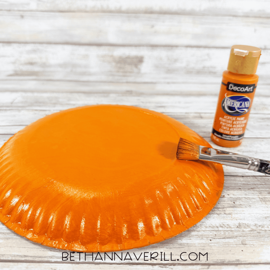 Paper plate painted bright orange with an orange paintbrush and bottle of orange acrylic paint beside it.