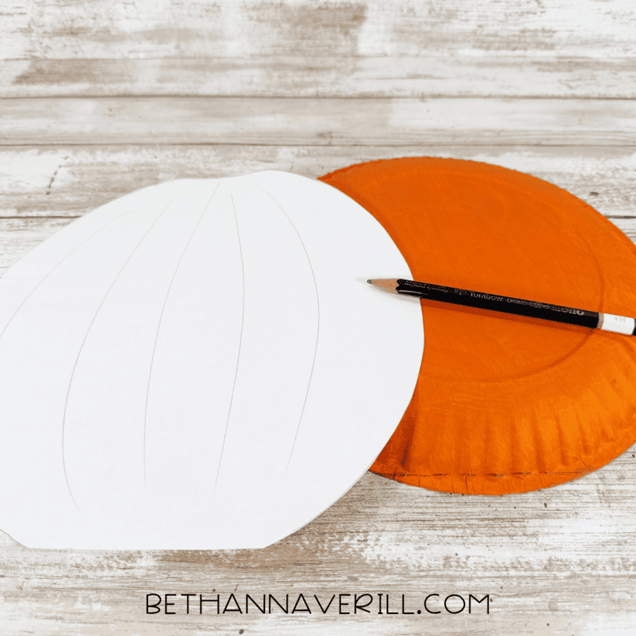 White paper pumpkin-shaped template beside an orange painted paper plate with a pencil resting across the plate.