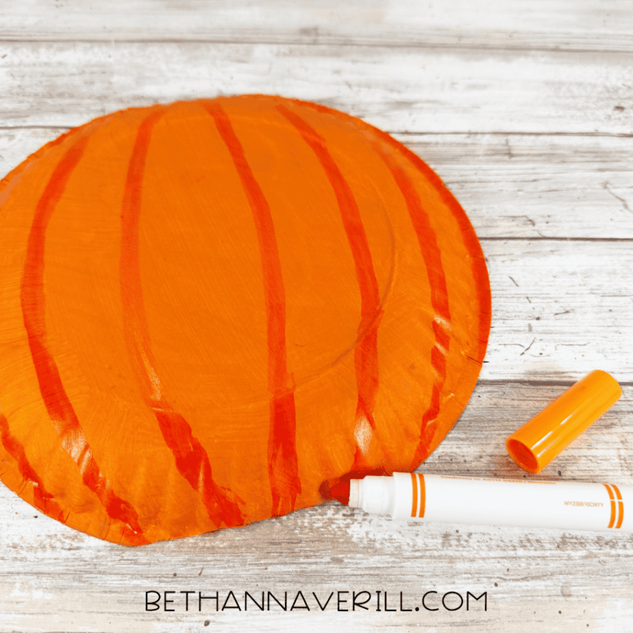Orange paper plate pumpkin with darker orange curved lines drawn from top to bottom and an orange marker beside it.