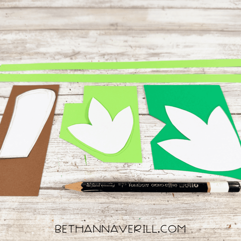 Green and brown paper pieces being traced from white templates, including two leaf shapes, a stem, and long green paper strips.