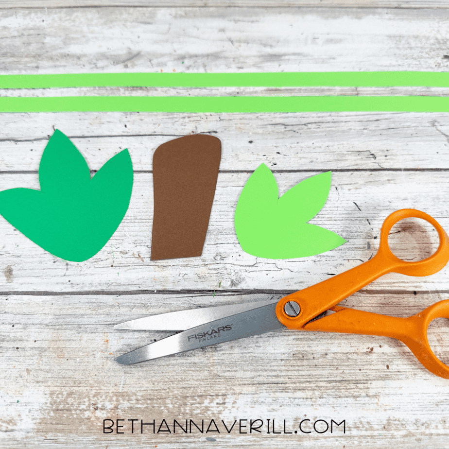 Cut green paper leaves, a brown paper stem, long green strips, and orange scissors arranged on a white wood surface.