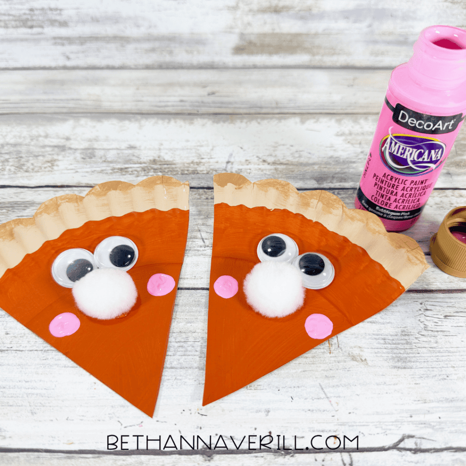 Two pumpkin pie paper plate slices with googly eyes, white pom pom noses, and pink painted cheeks.