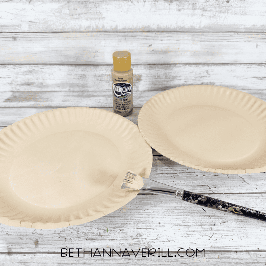 Two paper plates painted a light tan color with a paintbrush and bottle of acrylic paint nearby.