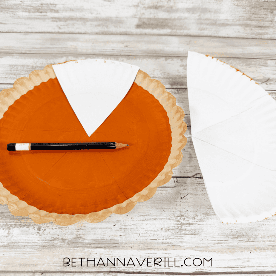 Orange paper plate divided into pie slice sections with pencil lines and a paper wedge used as a guide.