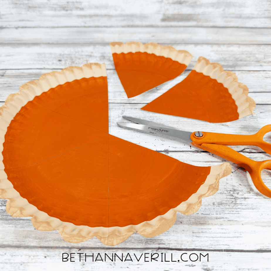 Pumpkin pie paper plate cut into several triangular slices with scissors on a white wood background.