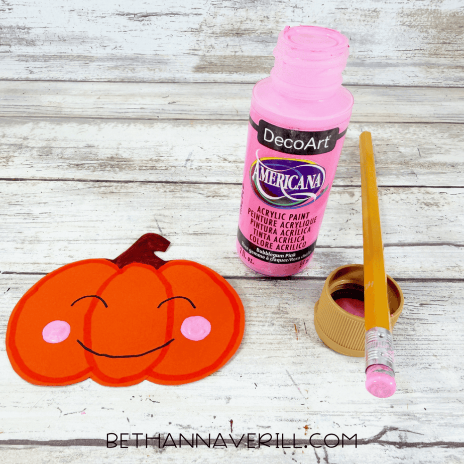 Orange pumpkin paper cutout with drawn face and pink cheeks beside an open bottle of pink paint and a pencil.