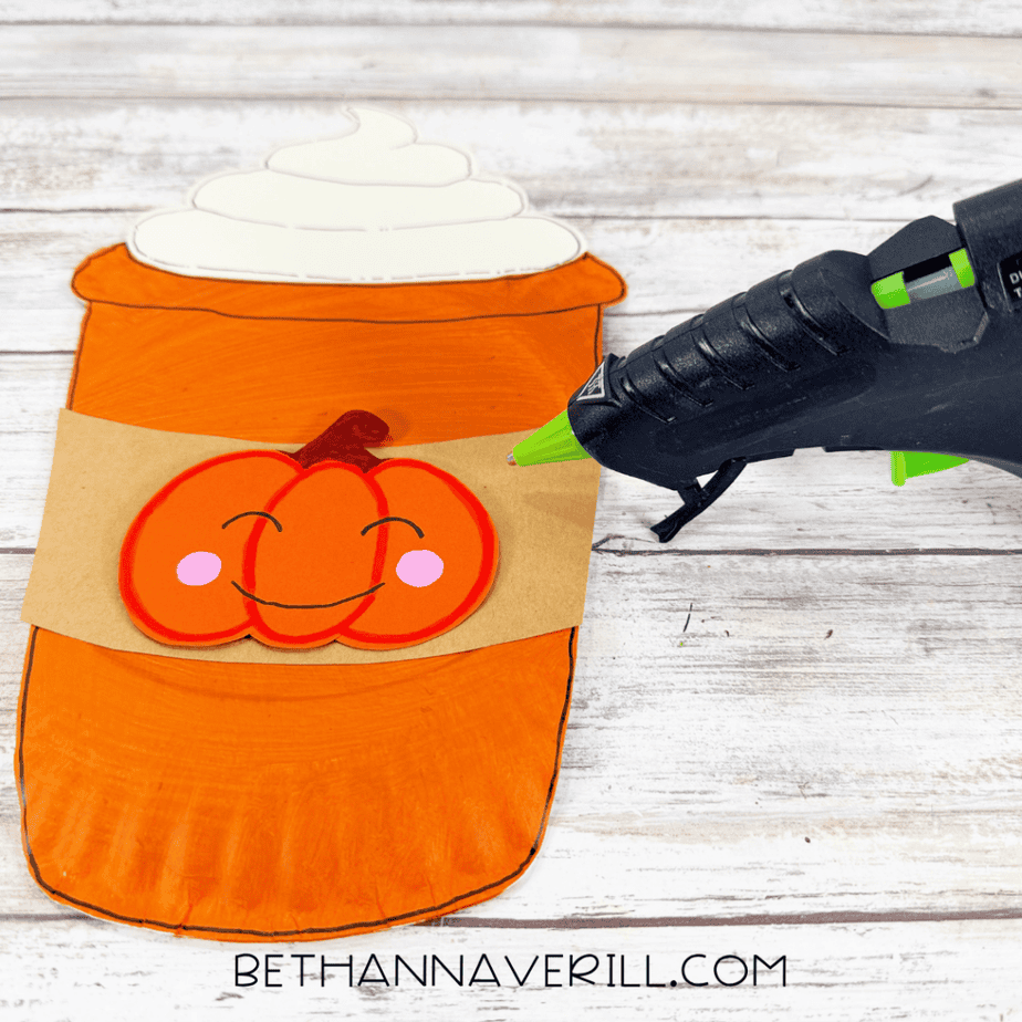 Pumpkin paper cutout attached to a brown sleeve on an orange paper plate cup with cream topping and a hot glue gun nearby.