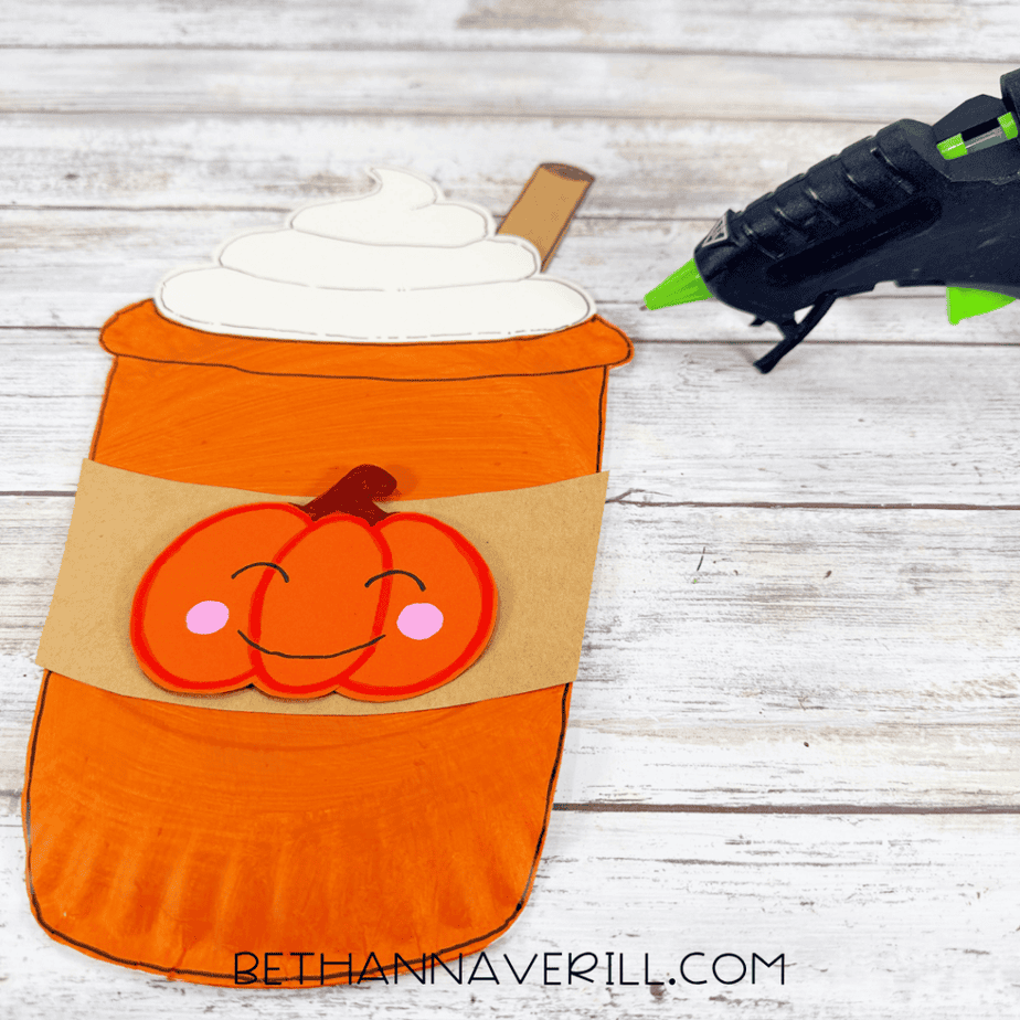 Orange paper plate cup with cream topping, brown stick, brown sleeve, and smiling pumpkin decoration assembled on the front.