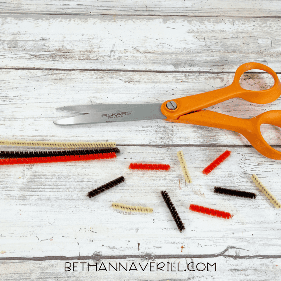 Orange, brown, and cream pipe cleaners cut into short pieces beside orange scissors.