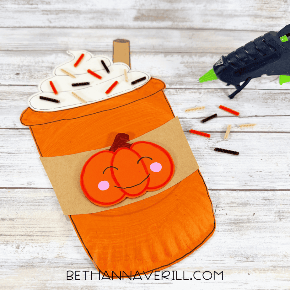 Finished orange paper plate cup craft with cream topping, short pipe cleaner pieces, brown stick, brown sleeve, and smiling pumpkin decoration.