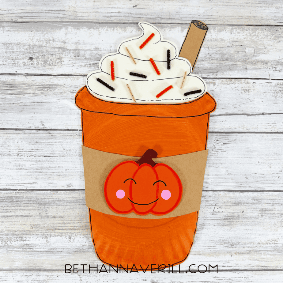 Finished orange paper plate cup craft with cream topping, brown stick, pipe cleaner pieces, and smiling pumpkin decoration on a white wood background.