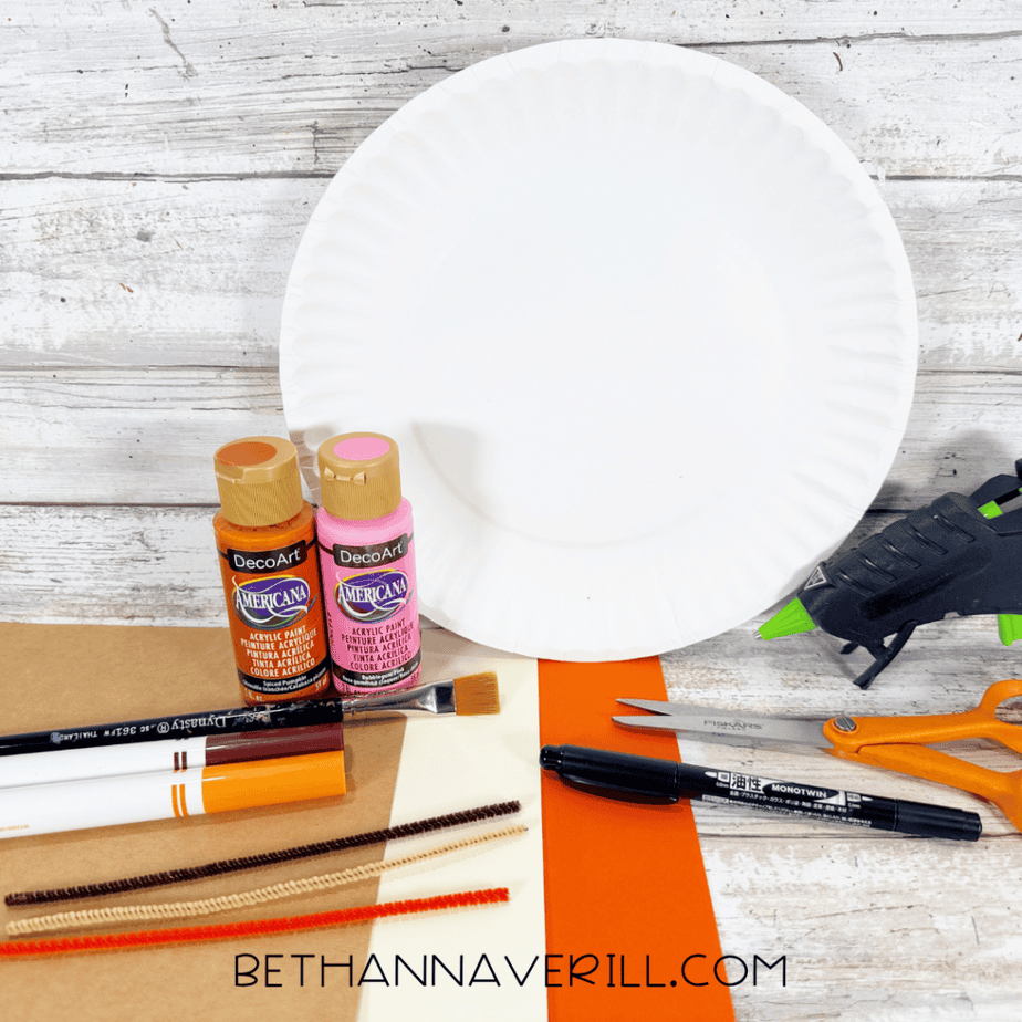 White paper plate with orange and pink acrylic paint, paintbrush, markers, pipe cleaners, scissors, and hot glue gun on a white wood surface.