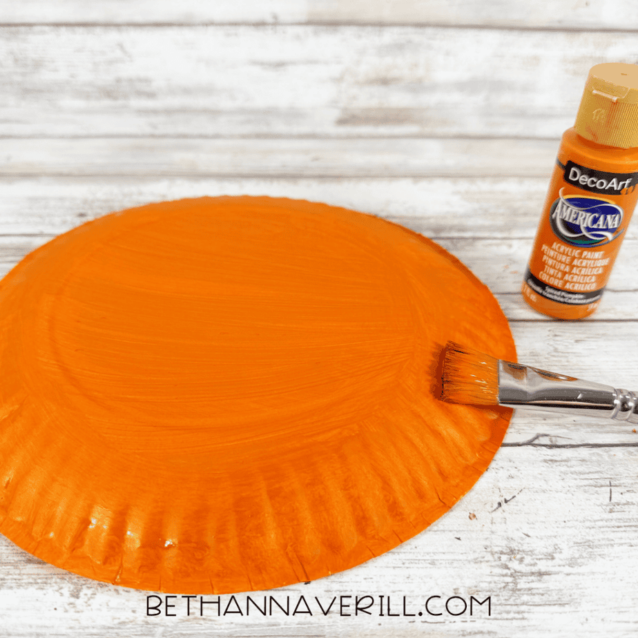 Paper plate painted orange with a paintbrush and bottle of orange acrylic paint beside it.