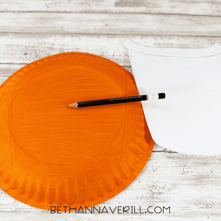 Orange painted paper plate with a curved rectangular shape traced in pencil and a white paper template beside it.
