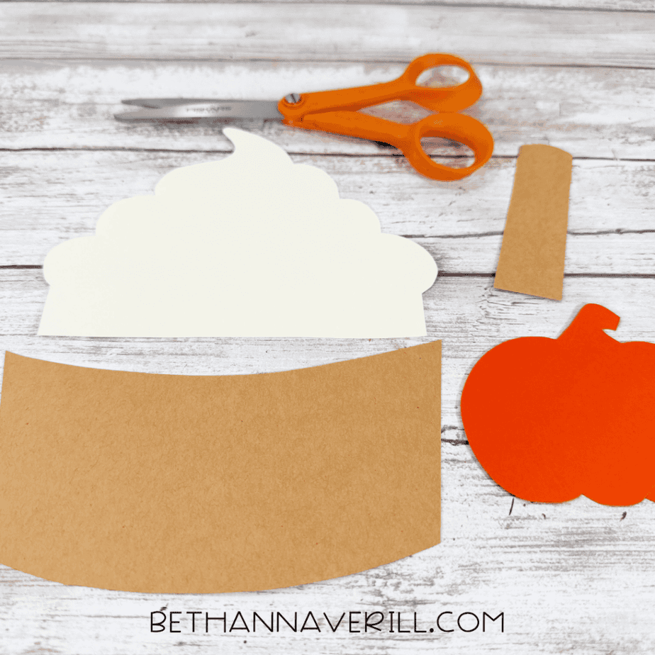 Cut paper pieces including a cream swirled shape, brown sleeve and stick, and orange pumpkin arranged with scissors.