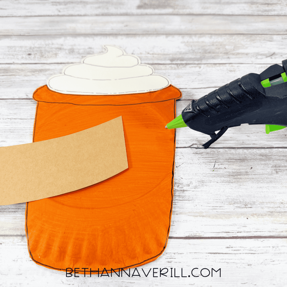 Orange paper plate cup with cream topping attached at the top while a brown paper sleeve is positioned across the front.